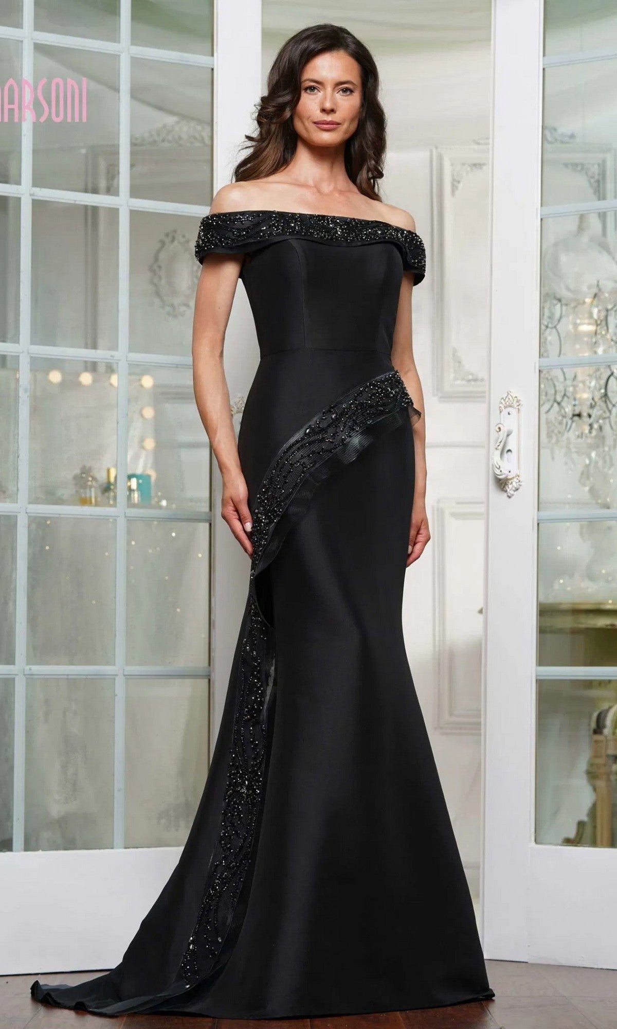 A woman stands indoors in front of glass doors wearing the Marsoni MV1332 long formal dress in black, featuring an off-shoulder mermaid silhouette with bead embellishments and crafted from Mikado fabric.